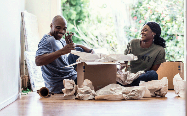 A military family sits on the floor of their new home while unpacking boxes.