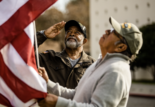 Two men standing in front of an American flag. One is raising the flag and the other is saluting the American flag.