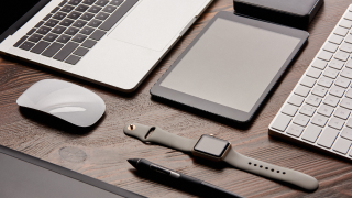 A desk with several electronic devices on it, including a laptop with mouse, an Apple Watch and a tablet